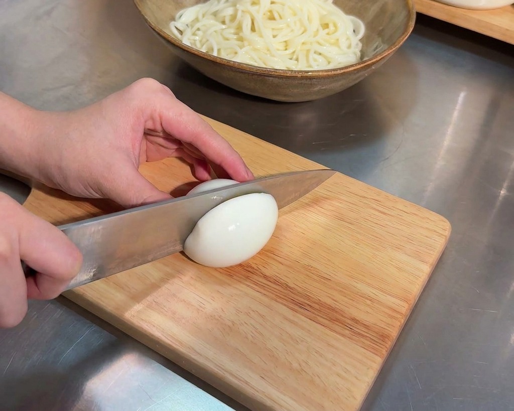 Un cuchillo de cocina afilado cortando por la mitad un huevo cocido sobre una tabla de madera clara.