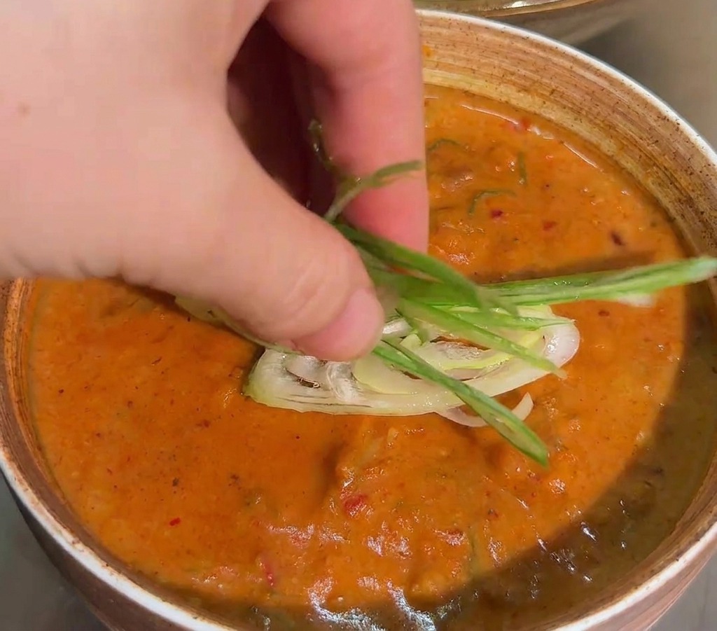 Una mano colocando tiras finas de cebolleta sobre la superficie de una salsa naranja espesa en un bol.