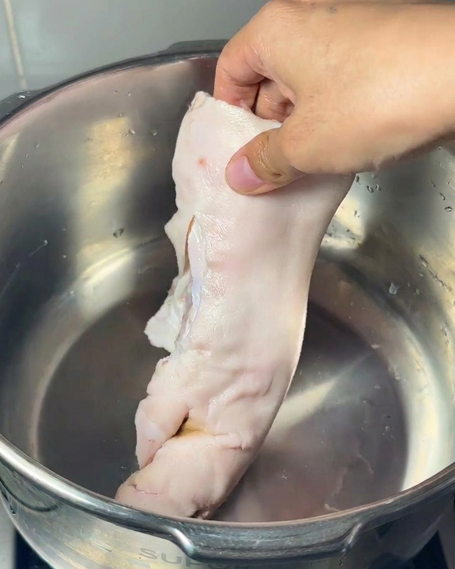 Mano colocando una manita de cerdo cruda en una olla de acero inoxidable junto a un hueso de caña grande y patas de pollo.