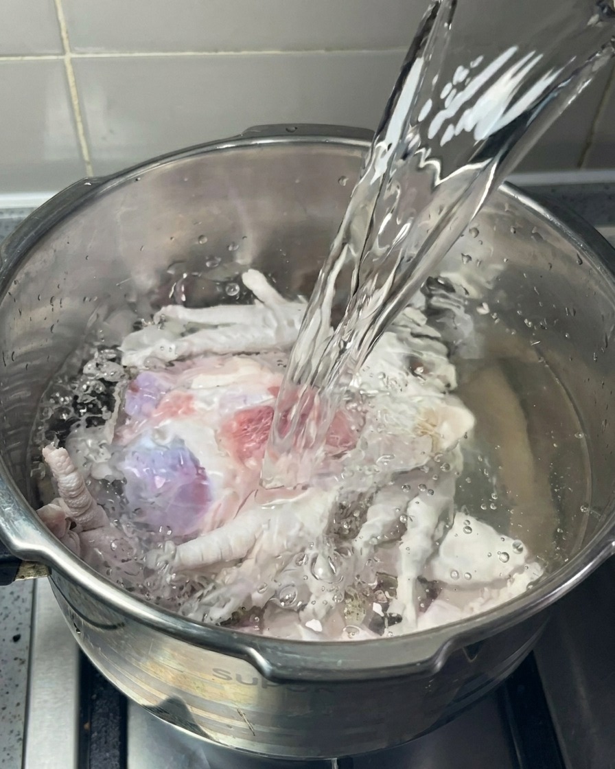 Chorro de agua clara cayendo en una olla de acero inoxidable llena de huesos de cerdo y patas de pollo.