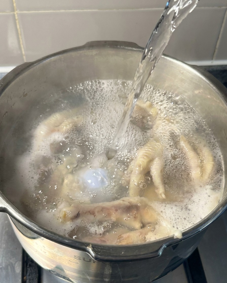 Agua hirviendo vertiéndose en una olla con patas de pollo y huesos de cerdo parcialmente cocinados.