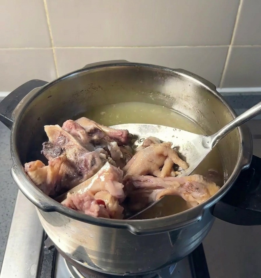 Colador de acero inoxidable levantando trozos de hueso de cerdo y patas de pollo cocidas de una olla con caldo amarillento.