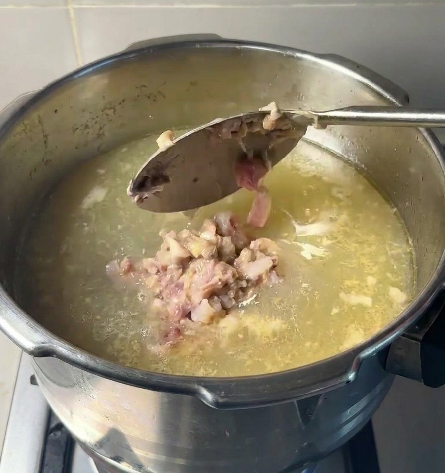 Cuchara metálica dejando caer trozos de carne machacada y tuétano en una olla con base de sopa clara.