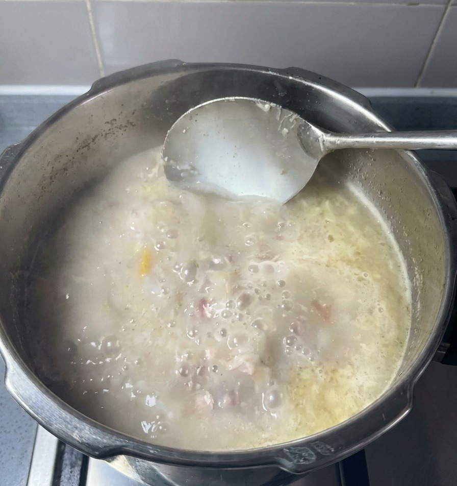 Cazo metálico removiendo una olla de caldo de huesos de cerdo opaco y blanco con trozos de carne y grasa.