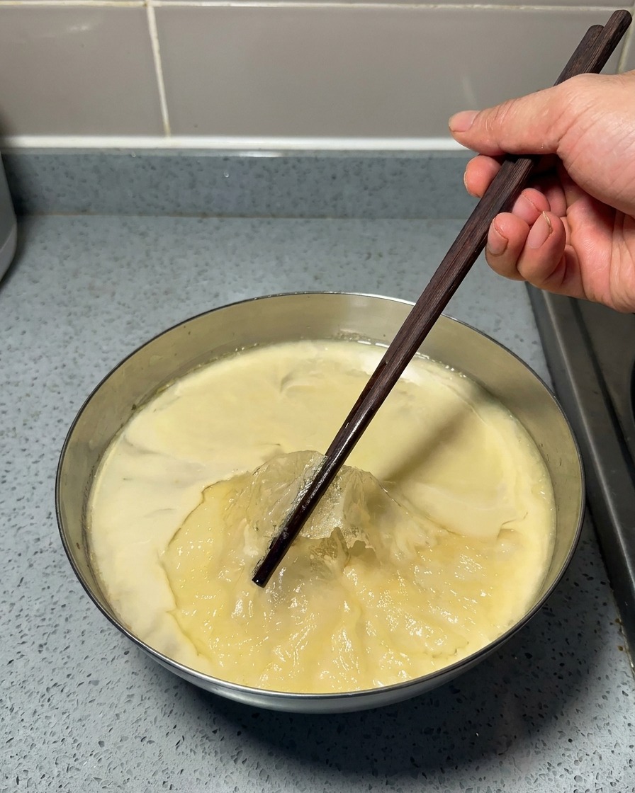 Mano usando palillos de madera para retirar una fina capa de grasa amarilla de la parte superior de un caldo gelatinoso frío.