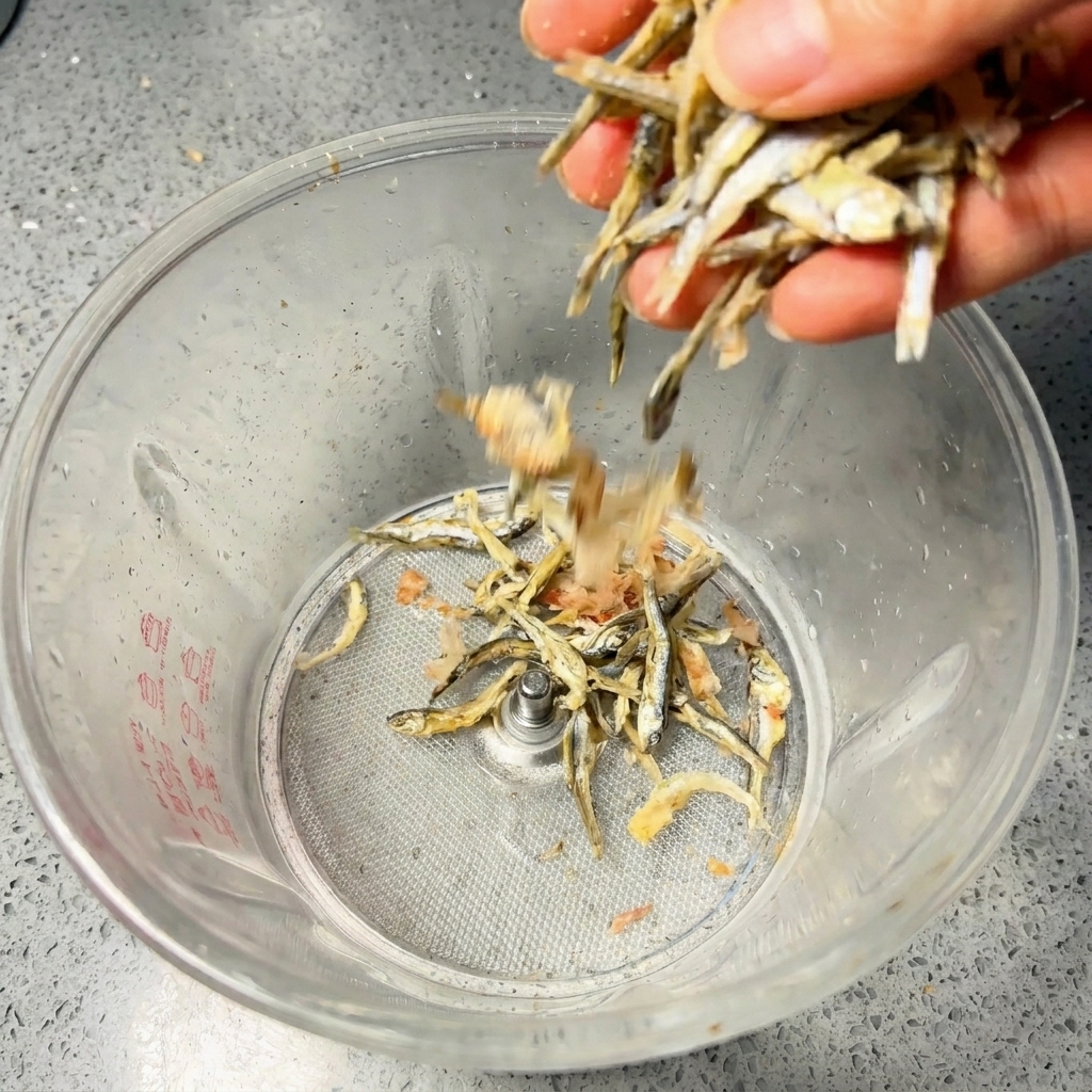 Una mano vertiendo anchovetas secas y copos de bonito en una licuadora