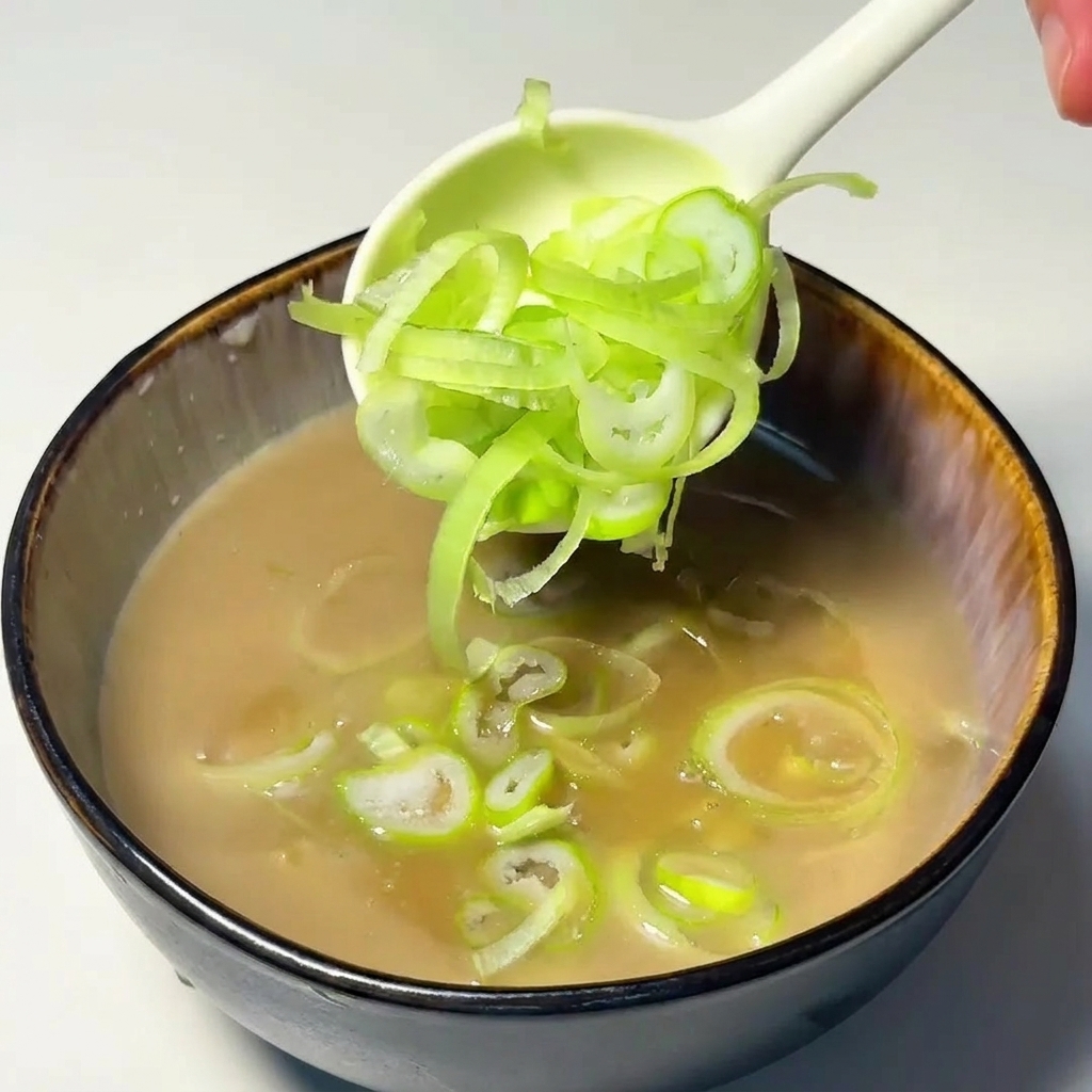 Una cuchara depositando cebolleta picada en la sopa de color canela