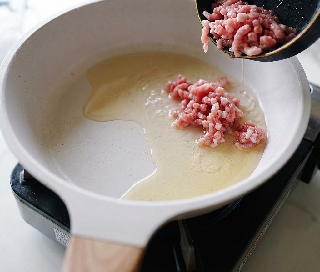 Carne de cerdo picada cruda vertiéndose en una sartén con aceite caliente.
