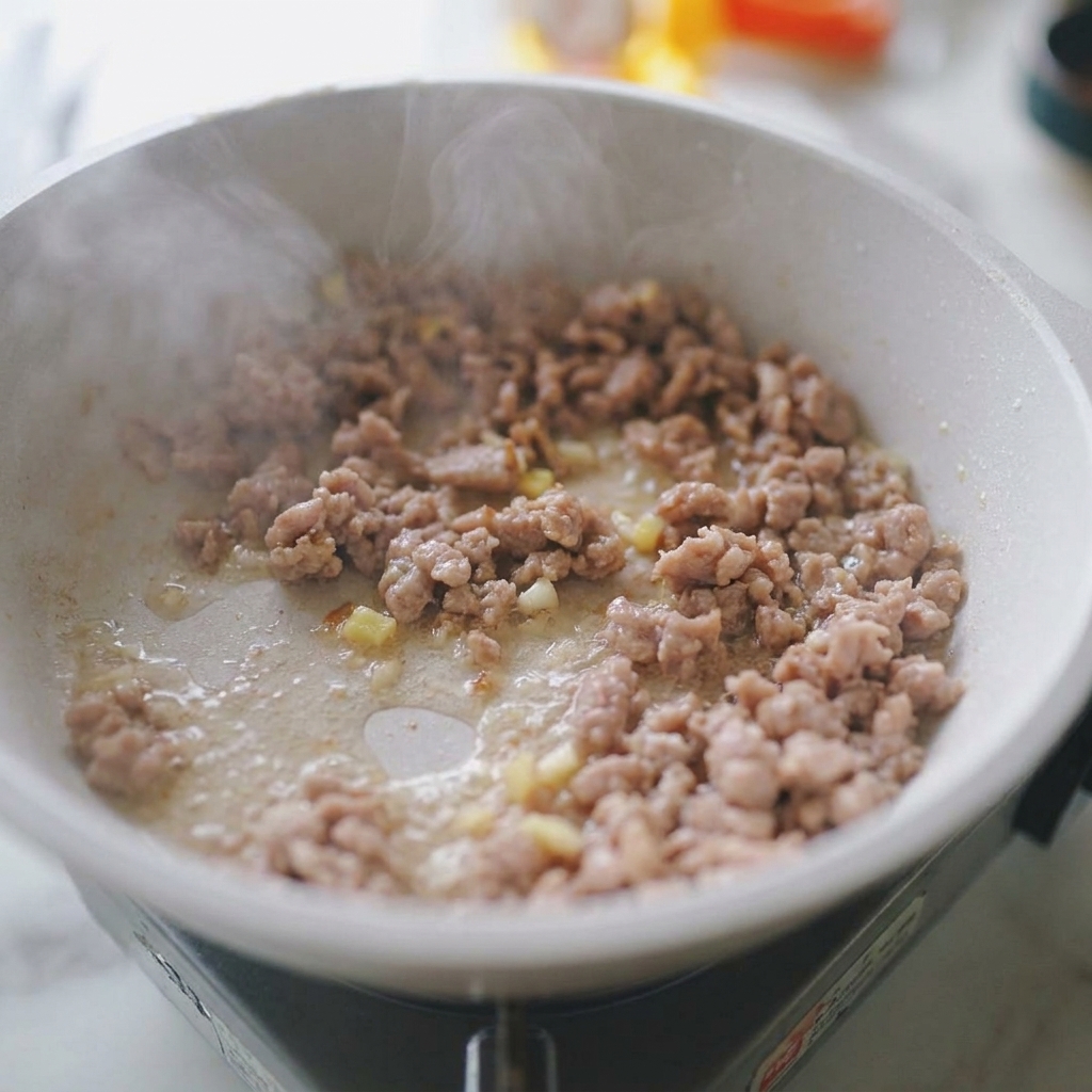 Carne de cerdo picada salteándose con trozos visibles de ajo y jengibre picado en una sartén.