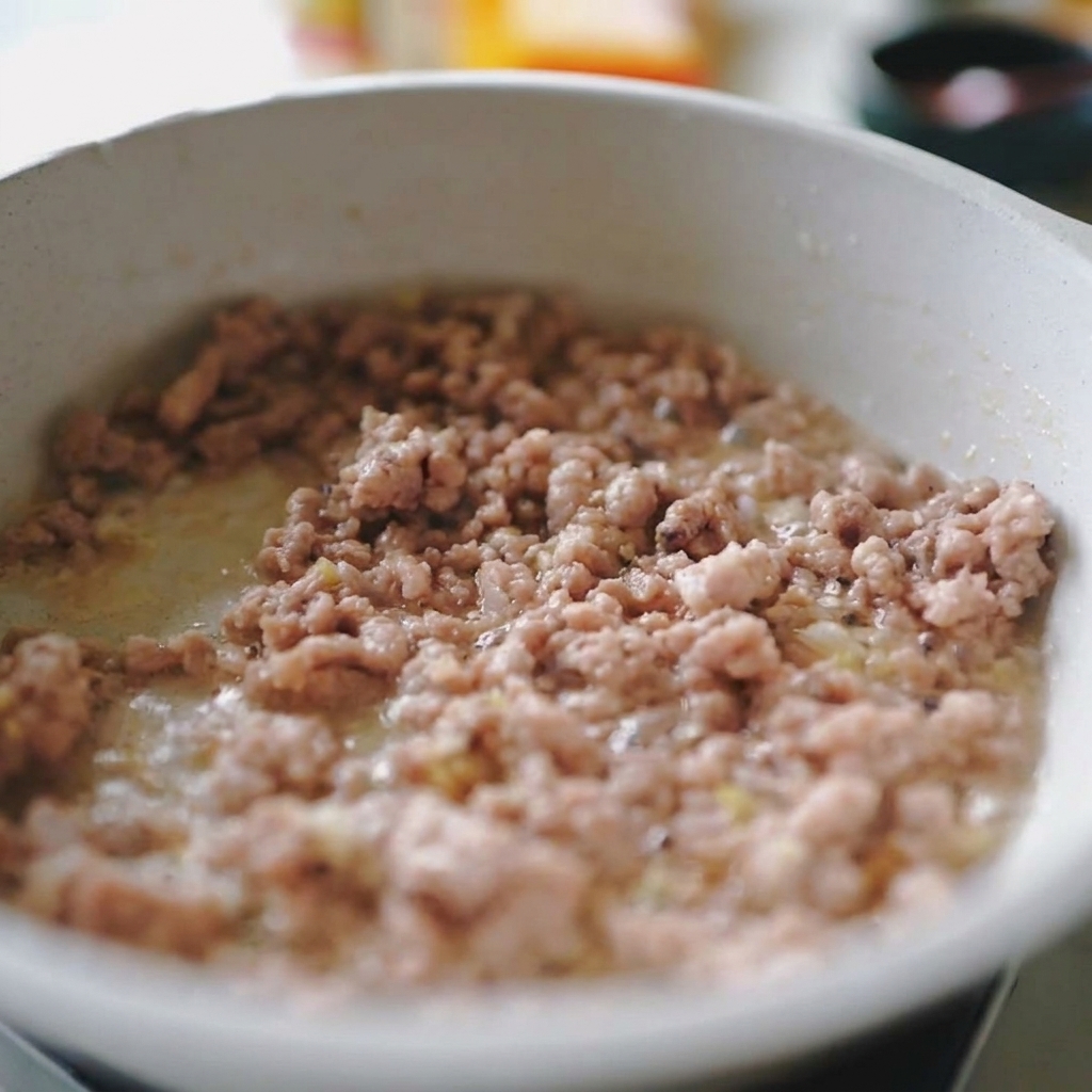Cerdo picado y tiras de cebolla blanca salteándose juntos en una sartén clara.