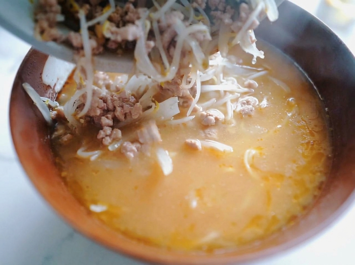 Primer plano de una cuchara añadiendo el cerdo salteado y los brotes de soja sobre el ramen en el bol.