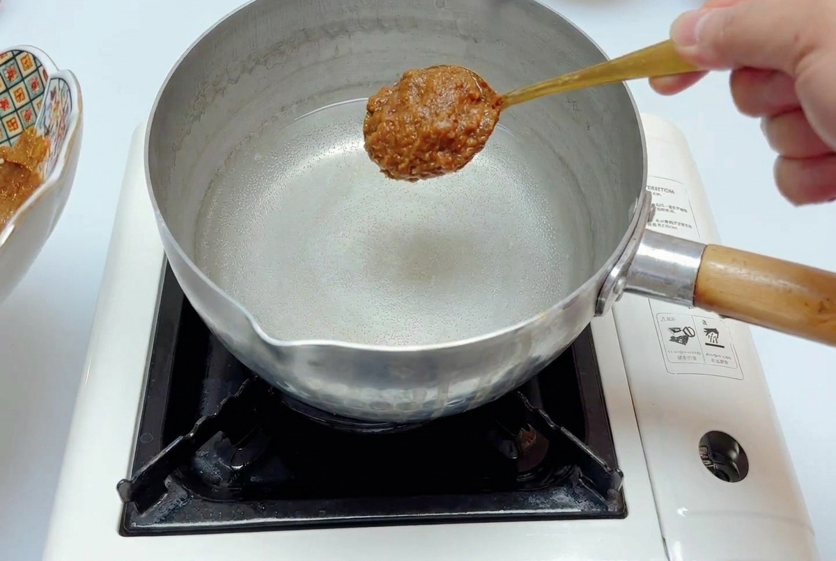 Cuchara con salsa de miso sobre una olla de acero inoxidable con agua en una estufa portátil.