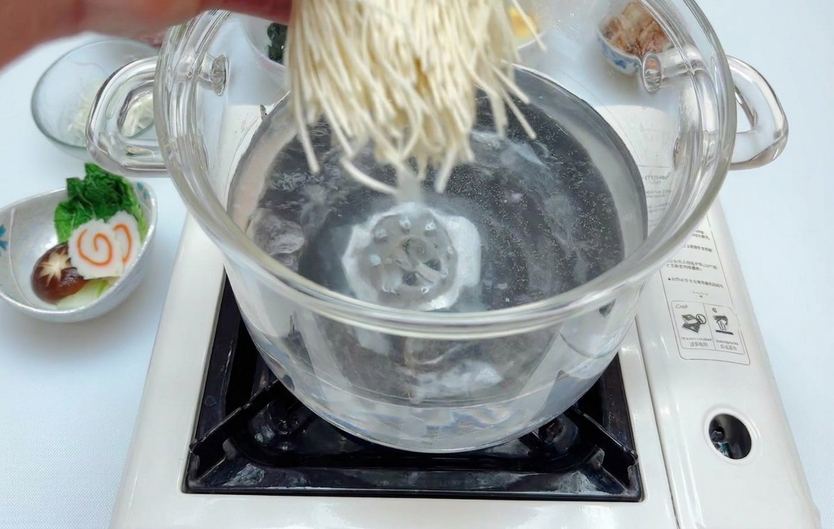 Fideos ramen cayendo en una olla de vidrio con agua hirviendo sobre una placa de inducción.