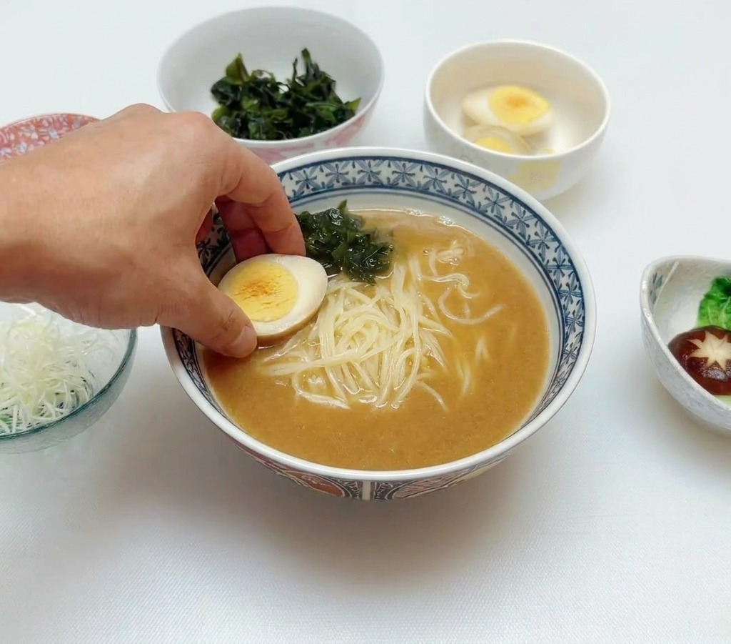 Mano colocando medio huevo mollet sobre el ramen que ya tiene caldo, fideos y alga wakame.