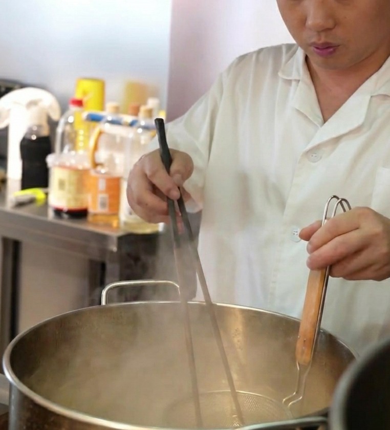Un chef bajando un colador de malla lleno de fideos frescos en una olla grande con agua hirviendo.