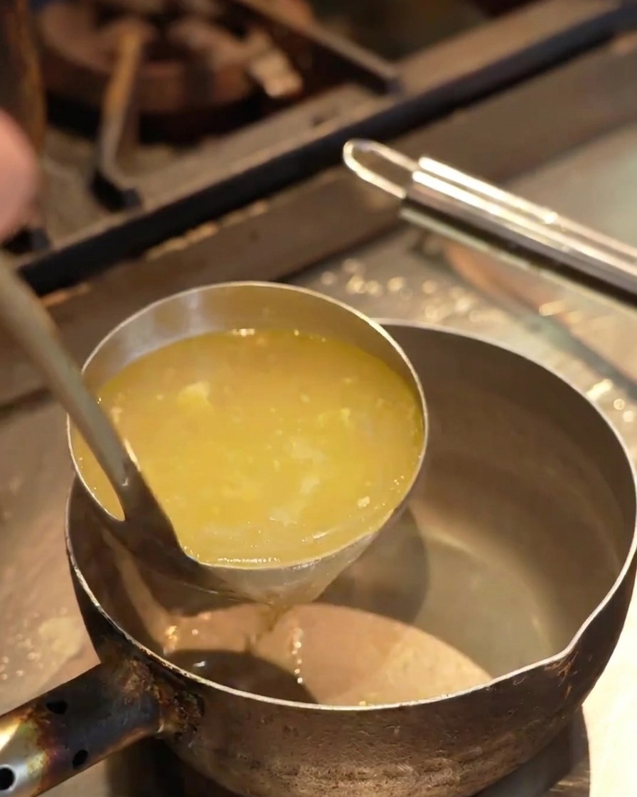 Caldo amarillo claro vertiéndose desde un cucharón en una pequeña olla de metal sobre una encimera de cocina.