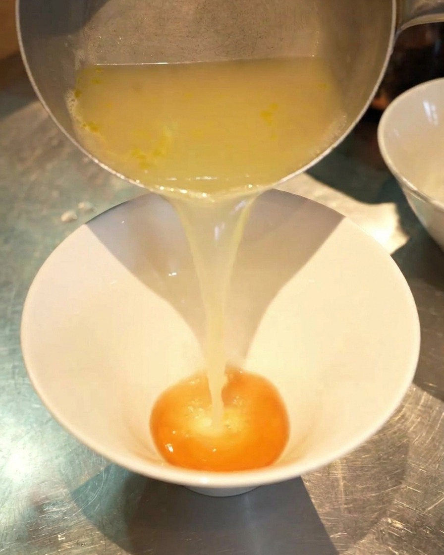Un chorro de caldo de pollo dorado y humeante vertiéndose desde una jarra plateada en un cuenco blanco con salsa naranja.