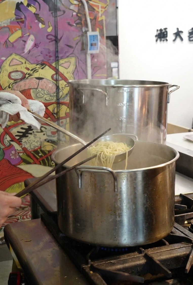 Una persona usando un colador para levantar fideos ramen cocidos de una olla grande de acero inoxidable con agua hirviendo.