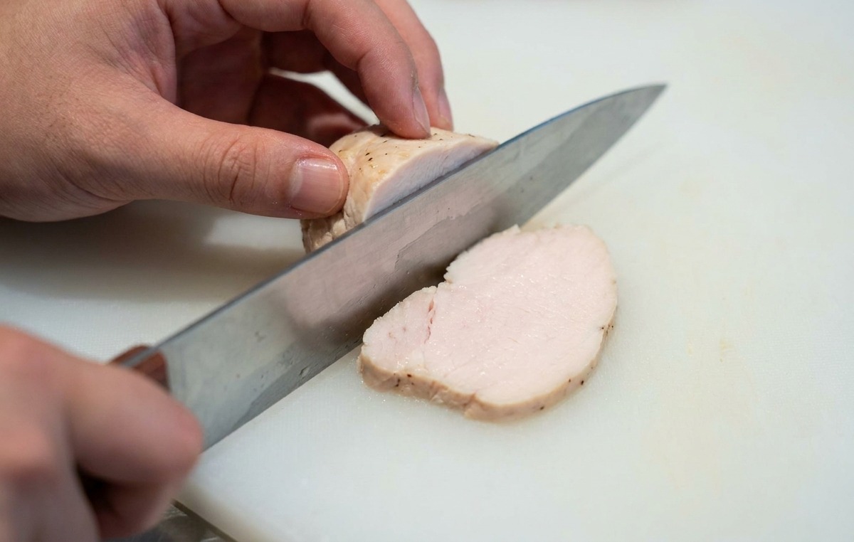 Manos de una persona cortando una pieza tierna de pechuga de pollo cocida en rodajas finas sobre una tabla de cortar blanca.