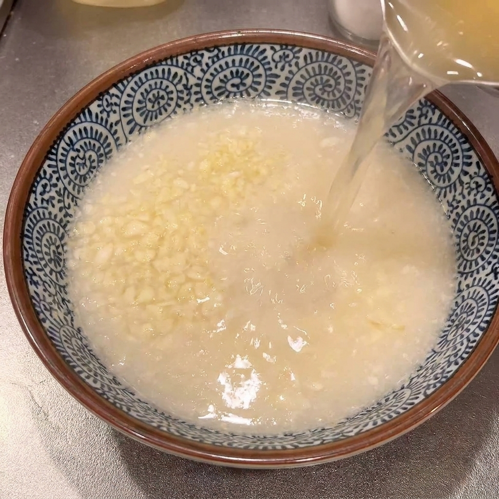 Caldo de ramen sazonado y caliente que se vierte desde una olla en un bol de cerámica decorado en azul y blanco que contiene ajo rallado.