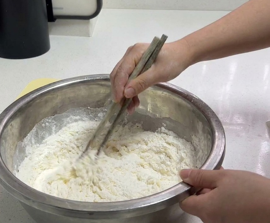 Una persona usando palillos de madera para mezclar harina de trigo blanca y agua en un bol grande de acero inoxidable.