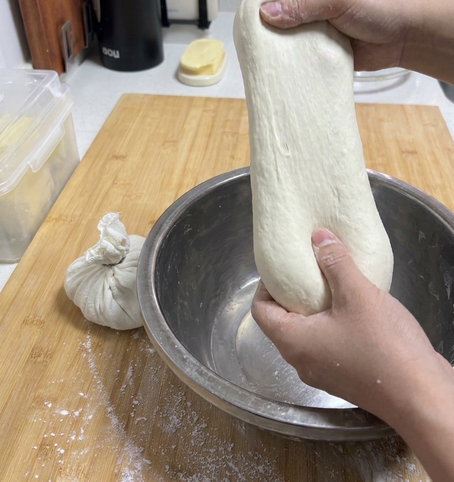 Manos de una persona estirando hacia arriba un trozo de masa pálida y suave desde un bol metálico para mostrar su elasticidad.