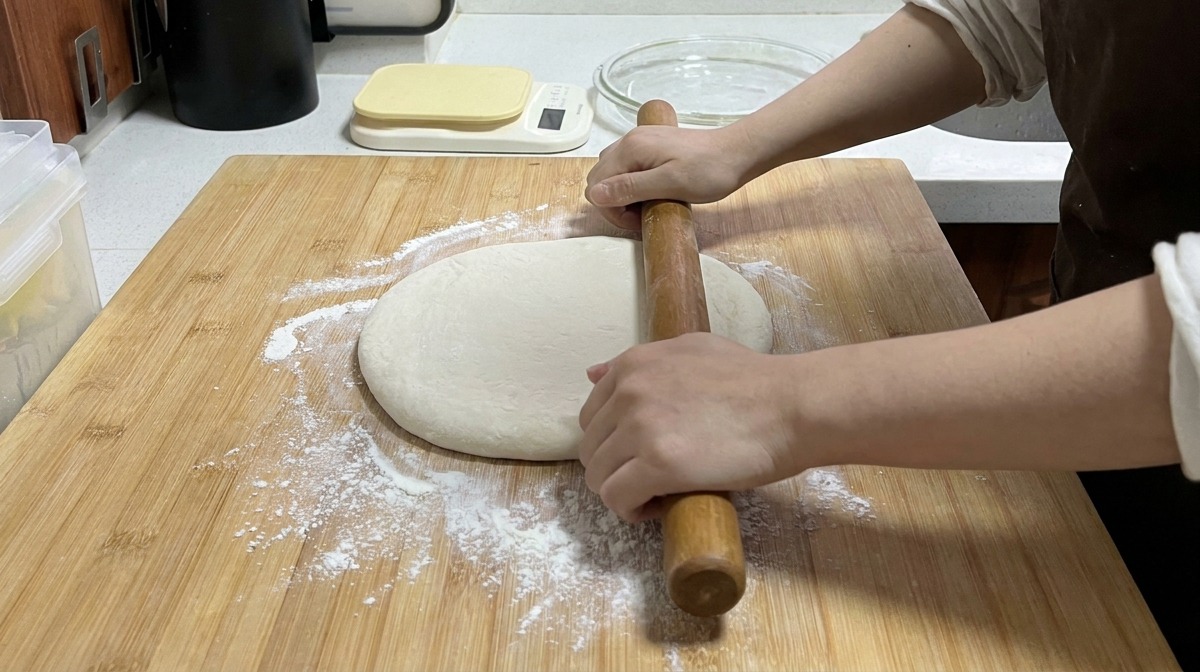 Manos usando un rodillo largo de madera para aplanar un trozo de masa sobre una tabla de bambú espolvoreada con harina.