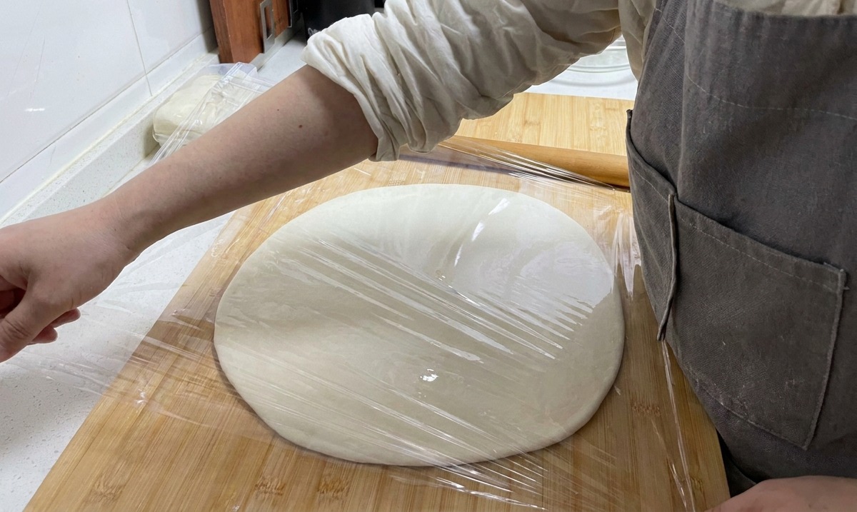 Una mano colocando film transparente sobre un círculo grande y plano de masa en una tabla de madera.