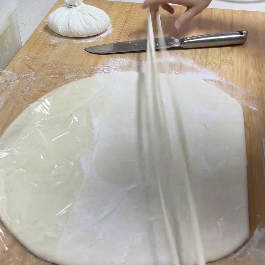 Una mano levantando una tira de masa de una tabla y estirándola para formar un fideo fino.