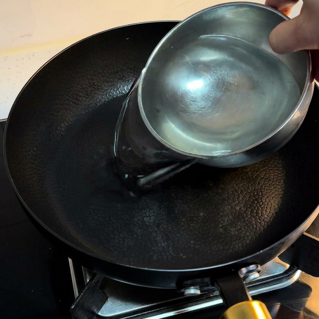 Agua vertiéndose desde un cuenco de acero inoxidable a una sartén negra en la cocina.