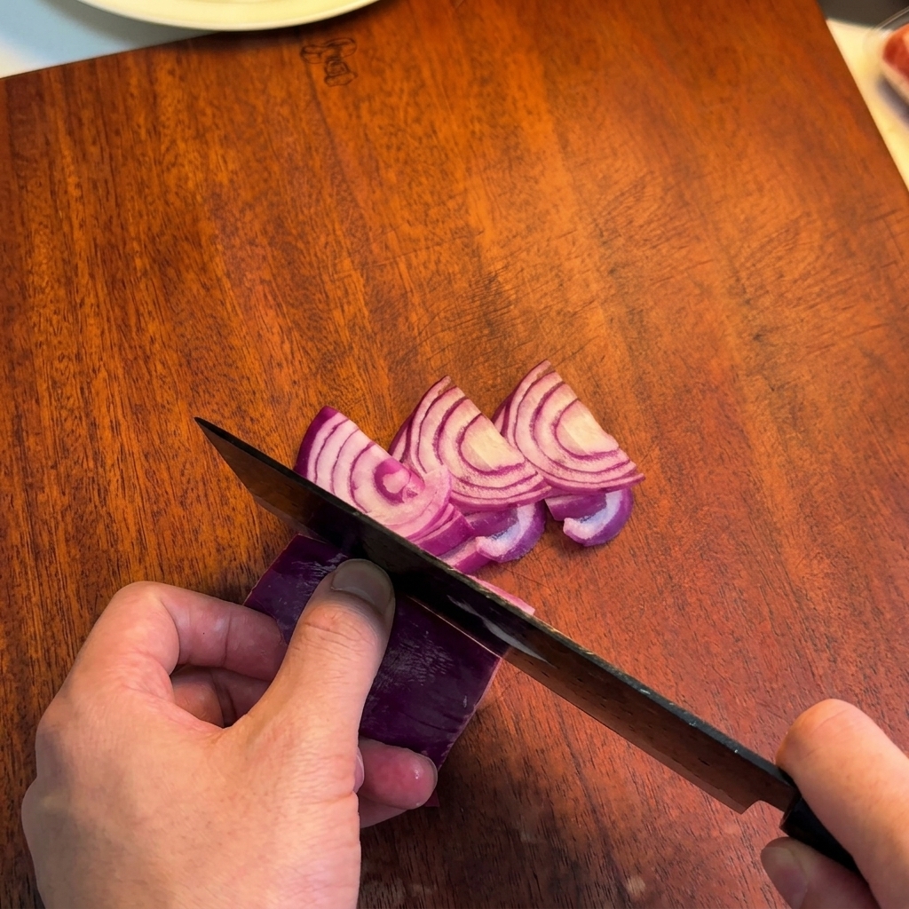 Una persona usando un cuchillo de chef para cortar una cebolla roja en tiras finas sobre una tabla de madera.