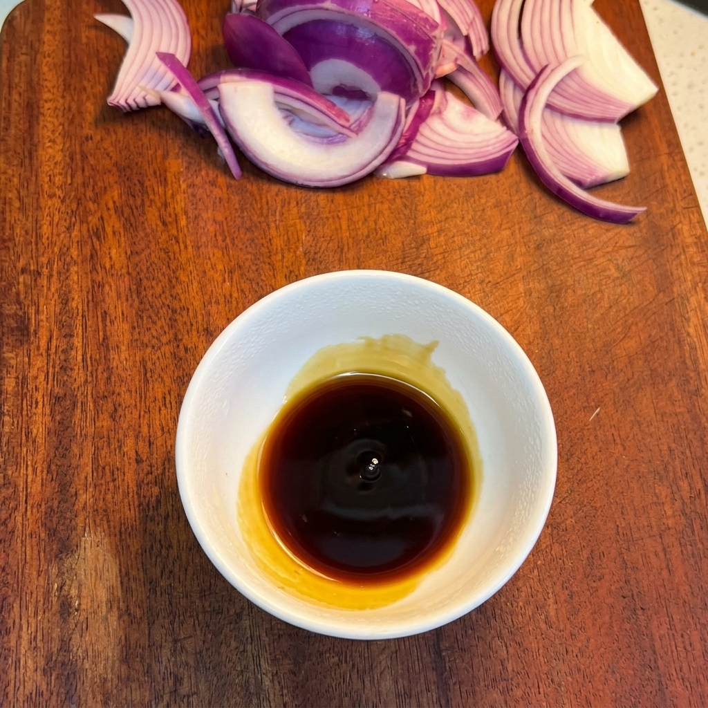Un cuenco blanco con una salsa oscura sobre una tabla de madera y cebollas al fondo.