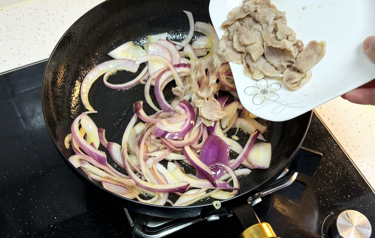 Láminas de ternera cocida vertiéndose desde un plato blanco a una sartén con cebollas salteadas.