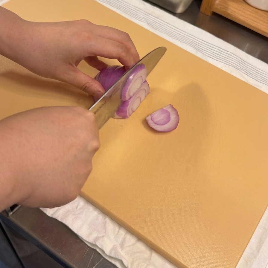 Manos cortando una cebolla morada en tiras finas sobre una tabla de madera.