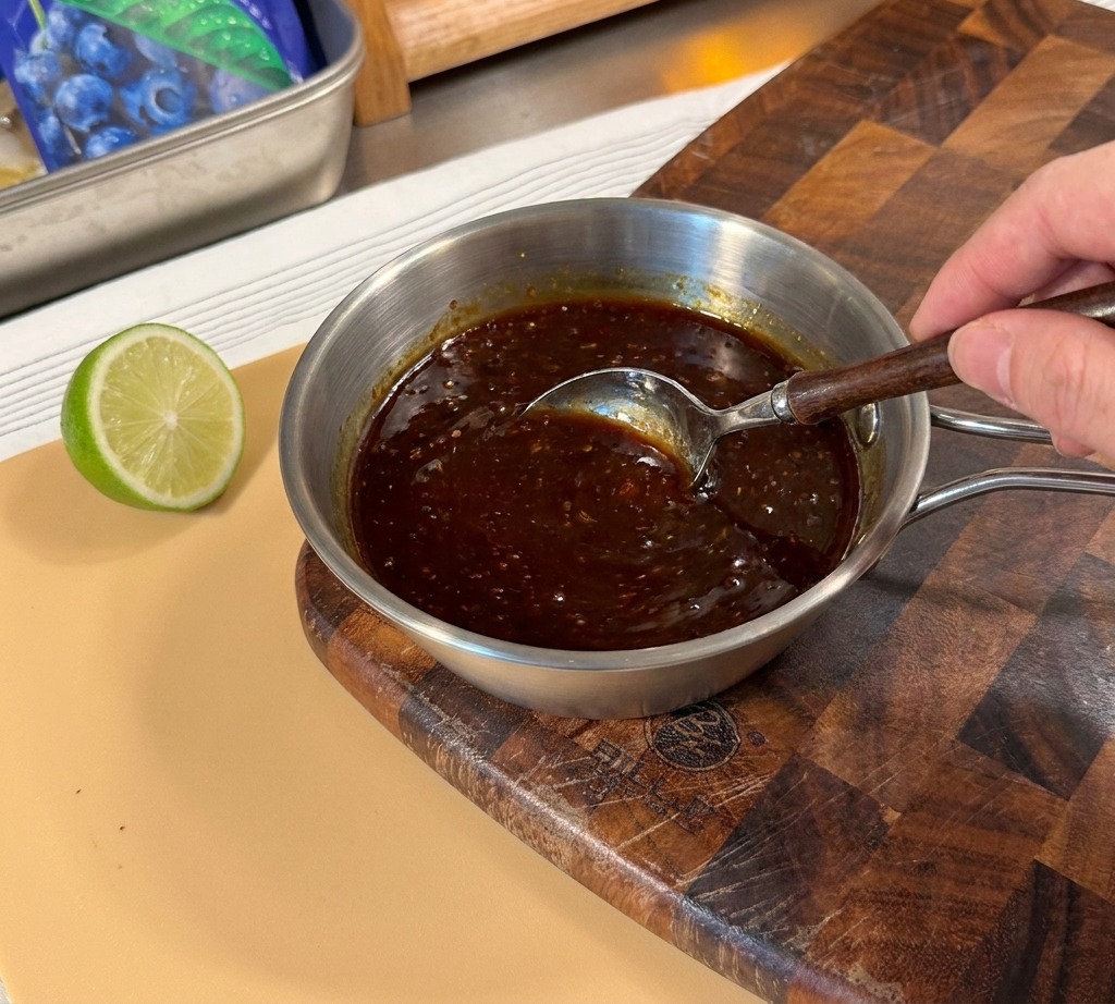 Una mano usando una cuchara para mezclar una salsa teriyaki oscura en un cazo de metal.
