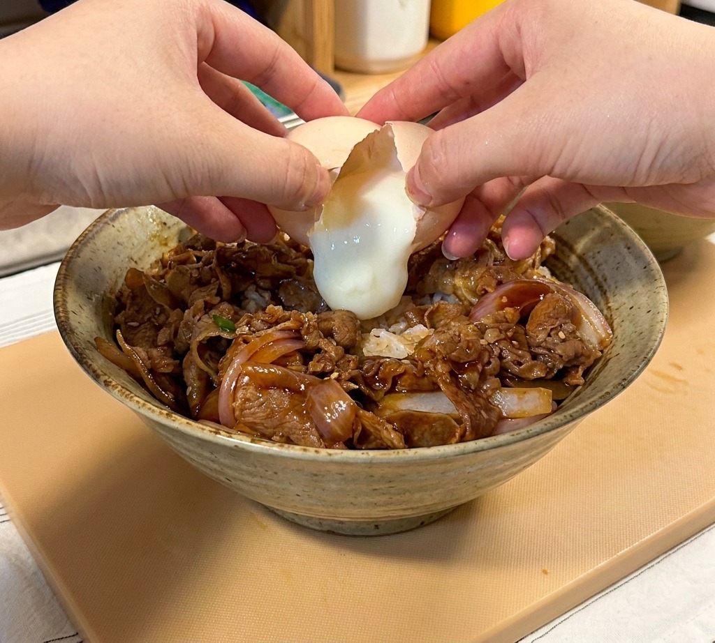 Dos manos rompiendo un huevo onsen poco hecho sobre un bol de ternera teriyaki y arroz.