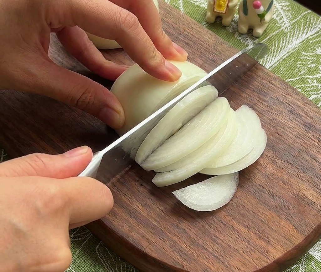 Manos de una persona usando un cuchillo de chef para cortar una cebolla blanca en tiras finas sobre una tabla de madera oscura.