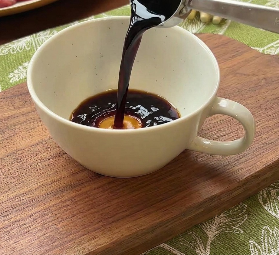 Una cuchara de metal vertiendo salsa de soja oscura en un bol de cerámica blanca con agua y una porción de pasta de chile coreana roja.