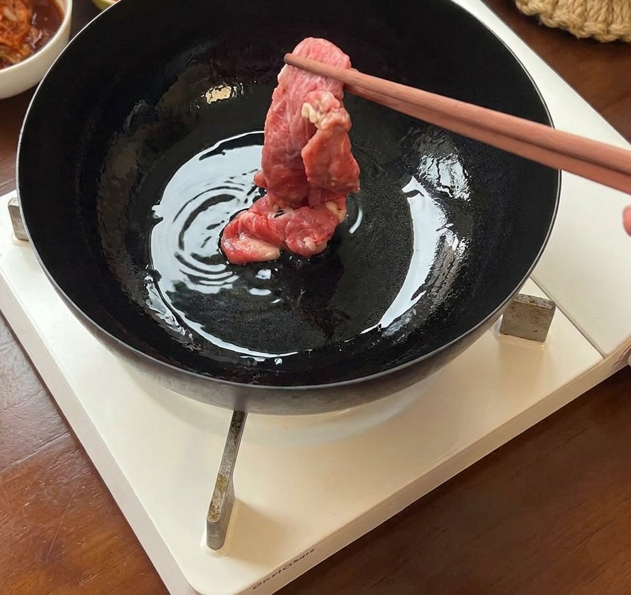 Láminas finas de ternera fresca cruda siendo colocadas en una sartén de hierro caliente con aceite usando palillos.