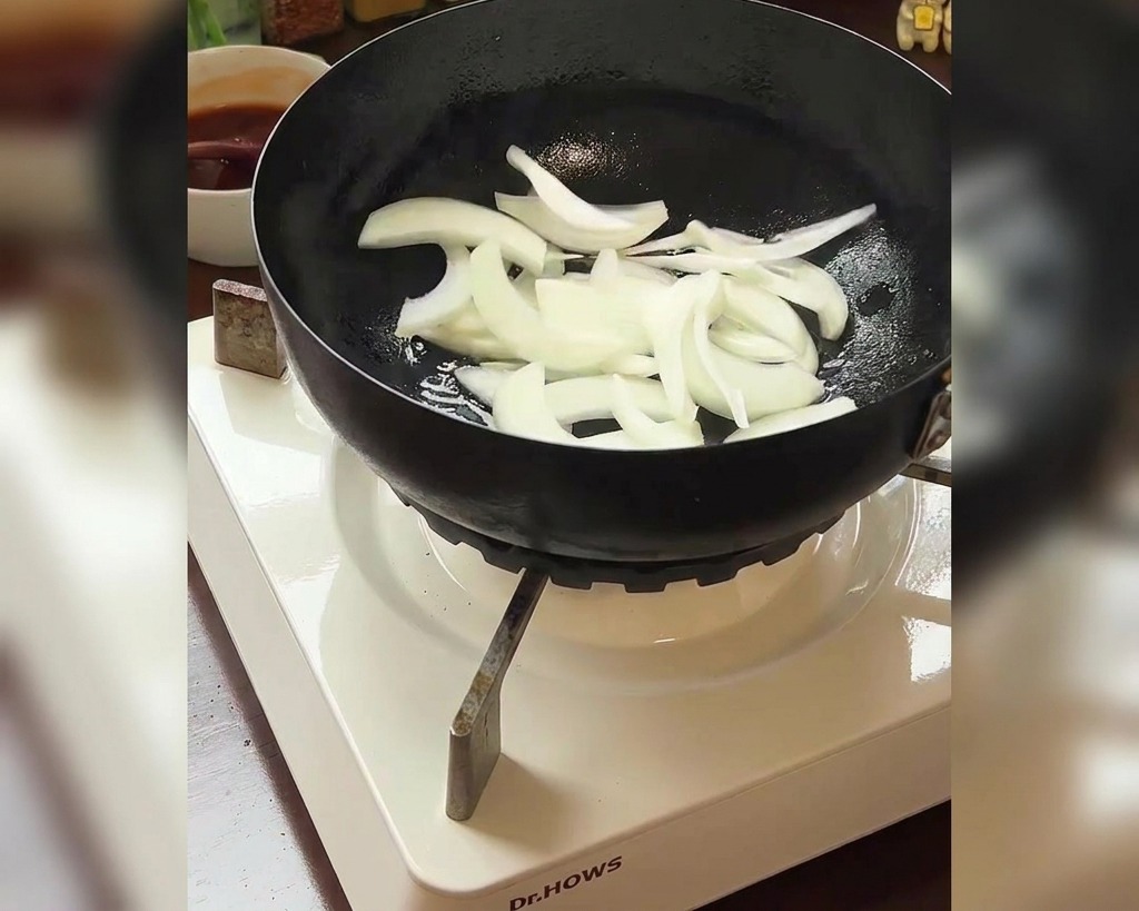 Tiras de cebolla blanca añadiéndose a una sartén con un bol de kimchi rojo visible al fondo.