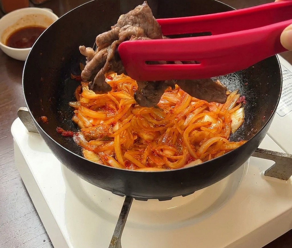 Láminas de ternera selladas volviendo a la sartén con el salteado de kimchi y cebolla usando unas pinzas rojas.