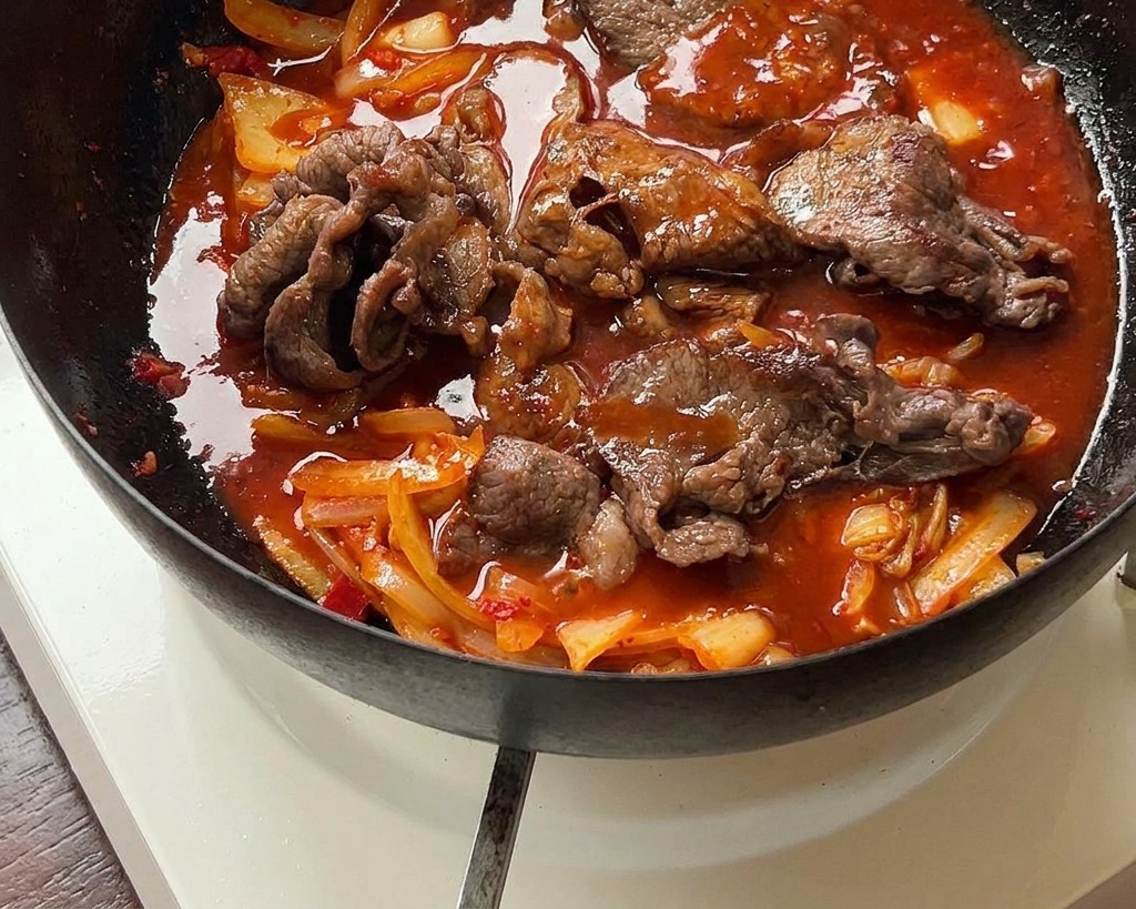 La mezcla de ternera y kimchi hirviendo a fuego lento en una salsa roja espesa dentro de una sartén de hierro negra.