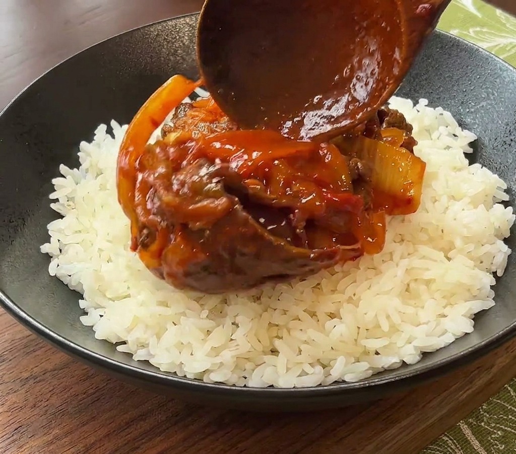 Una cuchara de madera sirviendo una porción de ternera y kimchi con salsa sobre una cama de arroz blanco en un bol de cerámica negro.