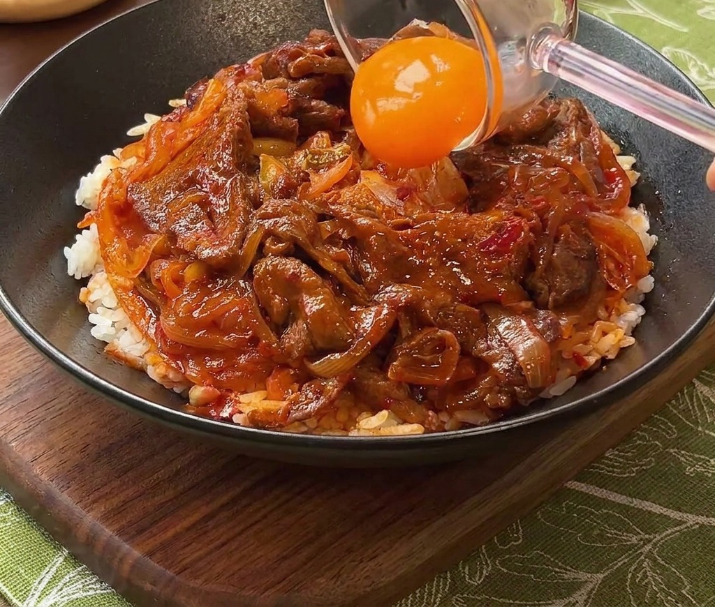 Una yema de huevo naranja brillante siendo colocada sobre el bol de ternera y arroz usando un separador de cristal.