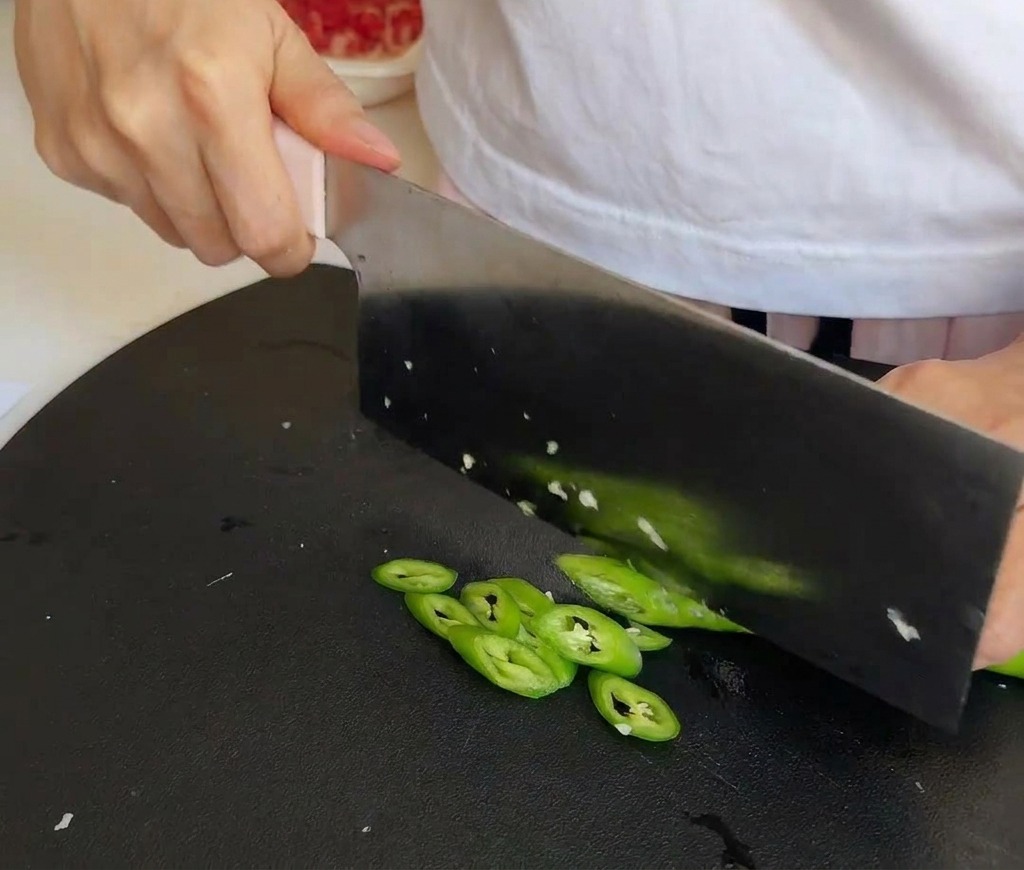 Un cuchillo de cocina cortando chiles verdes brillantes sobre una tabla de cortar negra.