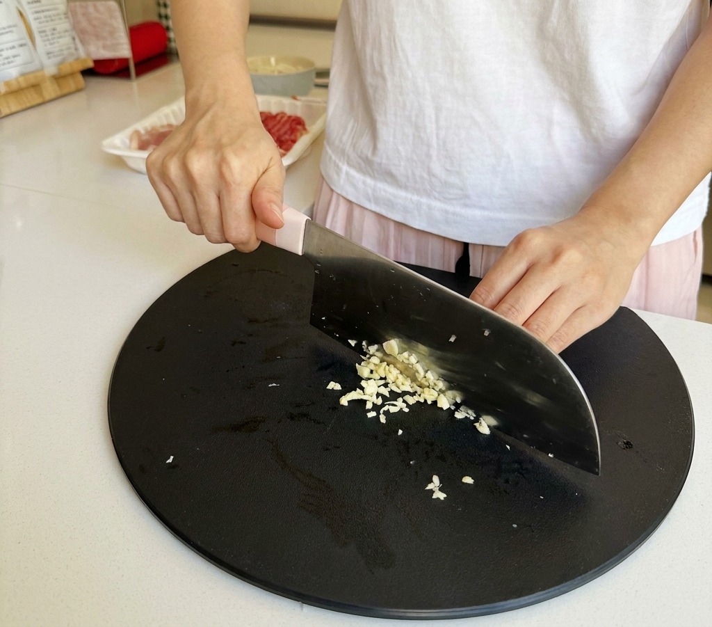 Primer plano de unas manos picando finamente dientes de ajo sobre una tabla de cortar negra.