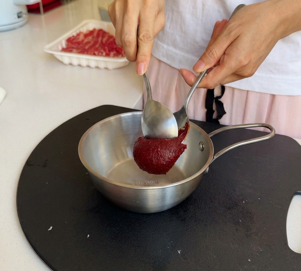 Una persona usando dos cucharas metálicas para añadir pasta de chile coreana roja a un bol de acero.