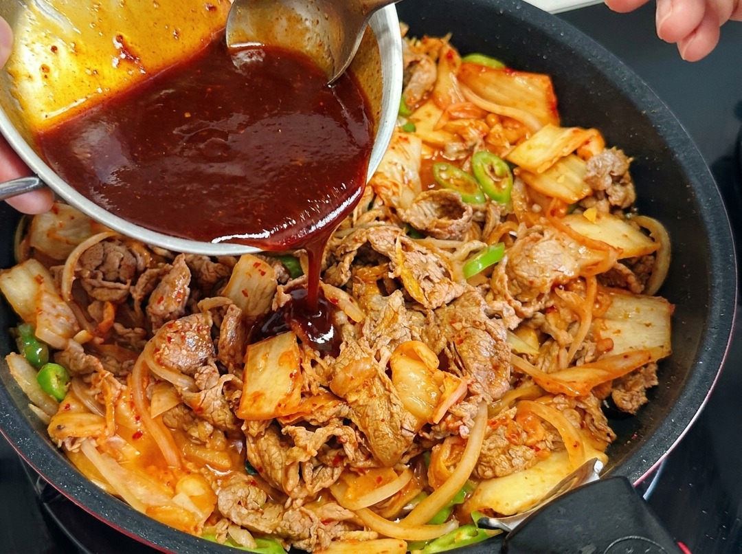 Una persona vertiendo una salsa roja desde un bol metálico a una sartén con ternera, cebolla, kimchi y chiles.