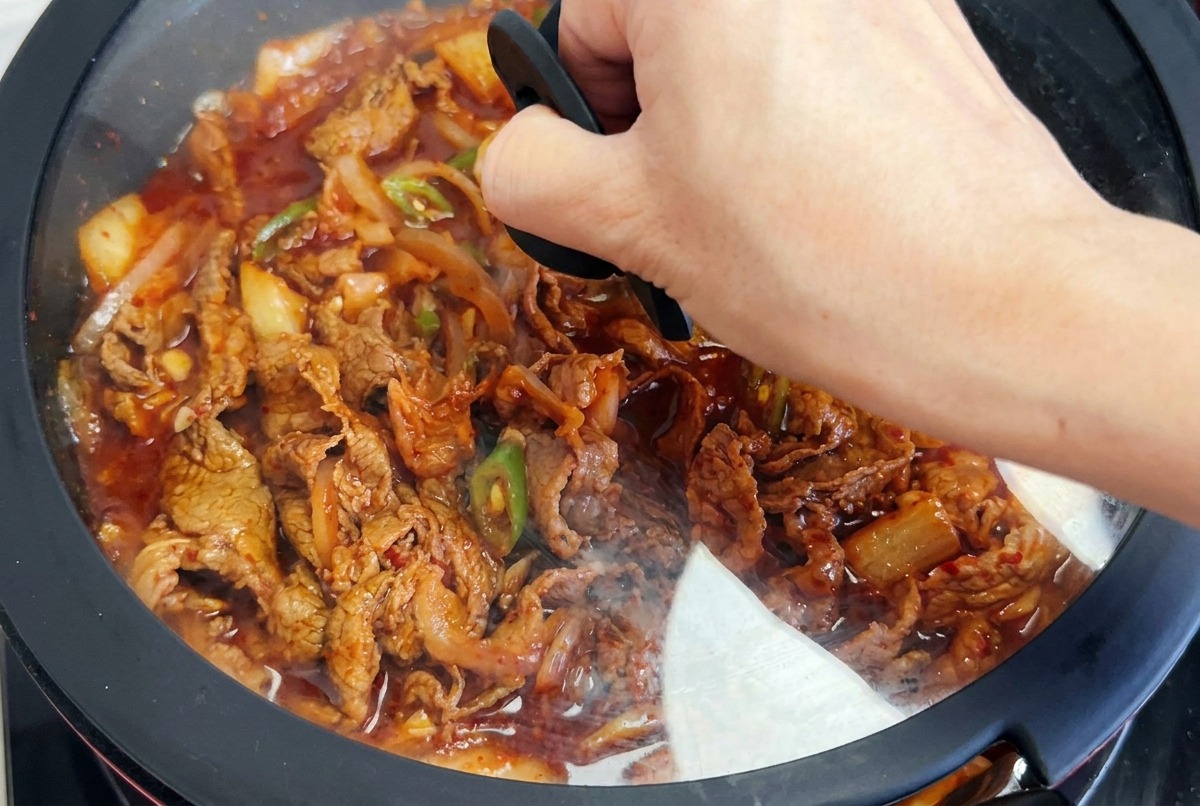 Una mano colocando una tapa de cristal sobre una sartén con ternera al kimchi y salsa roja hirviendo.