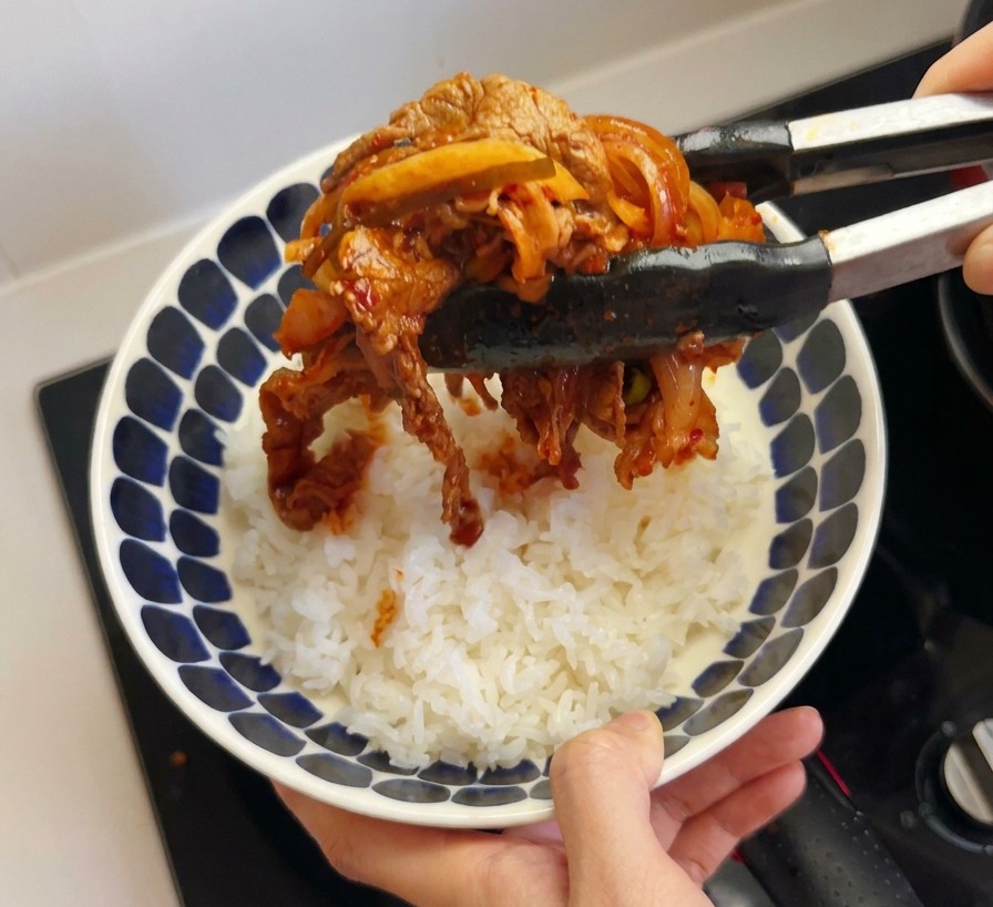 Unas pinzas de cocina colocando una ración de ternera y kimchi sobre un tazón de arroz blanco.